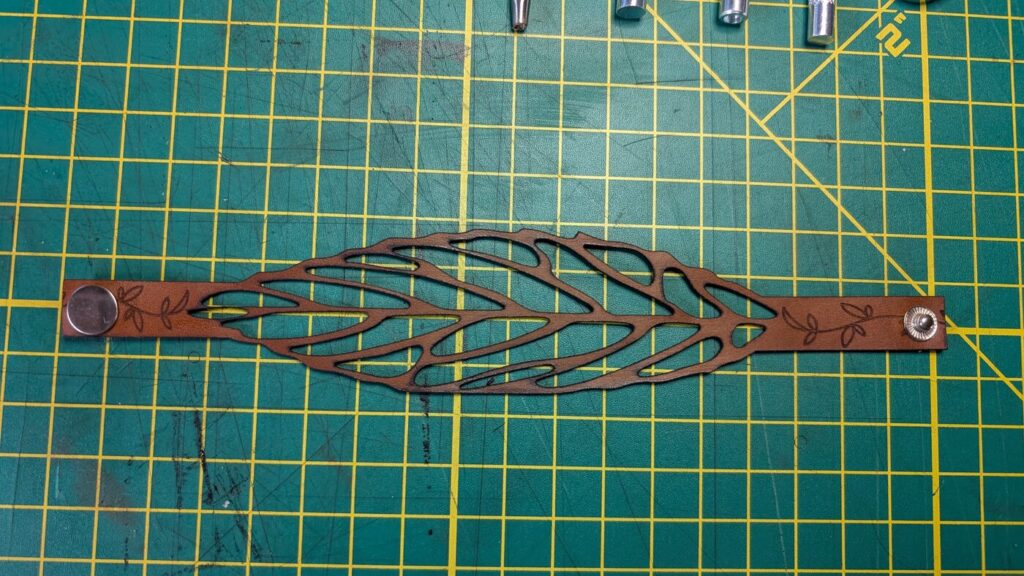 Laser-cut leather leaf cuff with snap closures on a green cutting mat in the Olsen Creations workshop