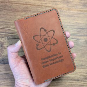 Tan leather Field Notes wallet laser-engraved with an atom and "Imagination is more important than knowledge", held in hand