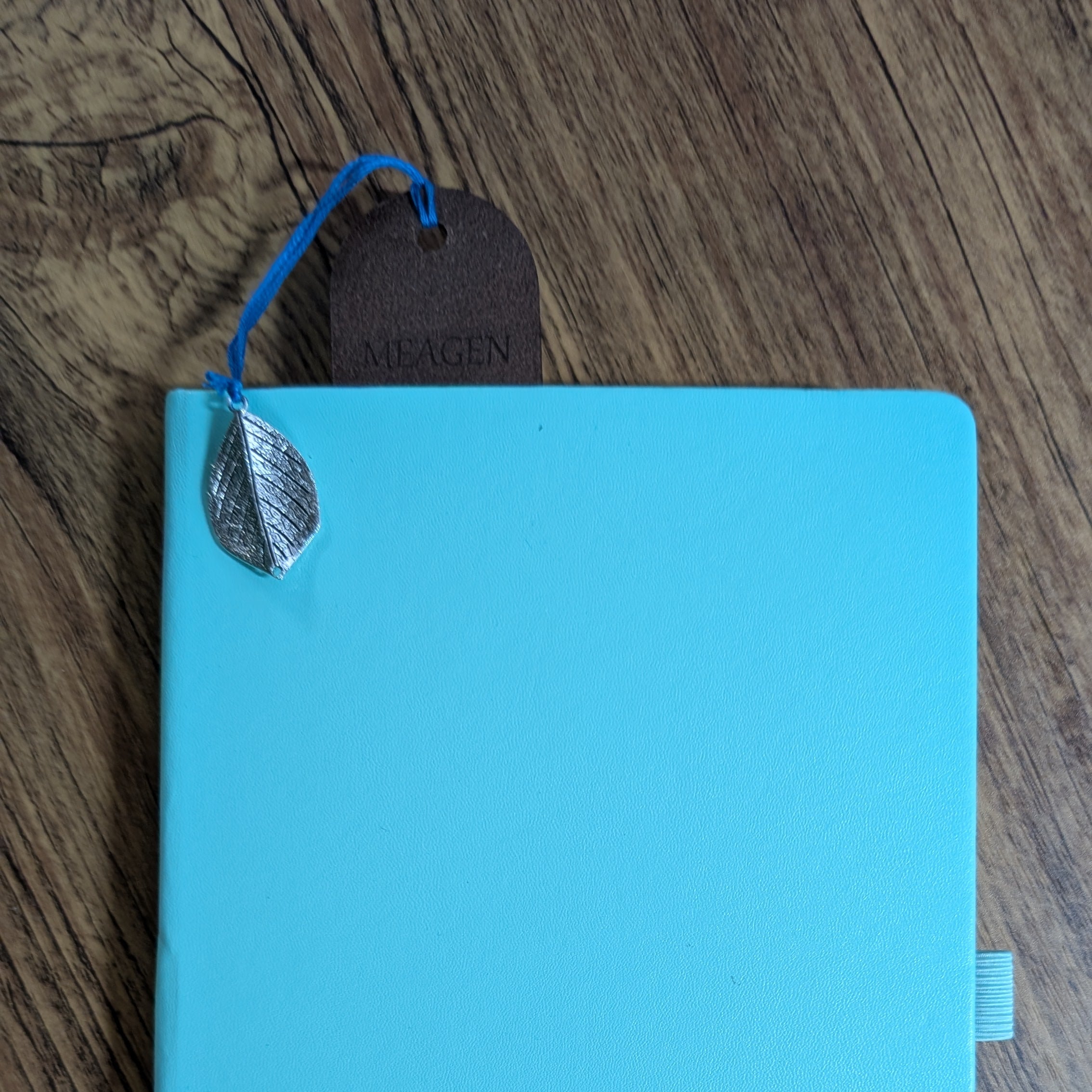 Personalized leather bookmark tucked into a teal book, with a silver leaf charm