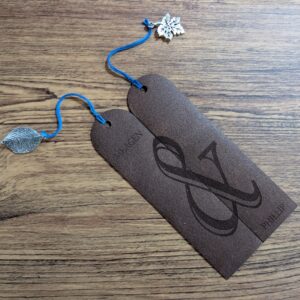 Two brown leather bookmarks engraved with names that join into one ampersand, with leaf and maple leaf charms